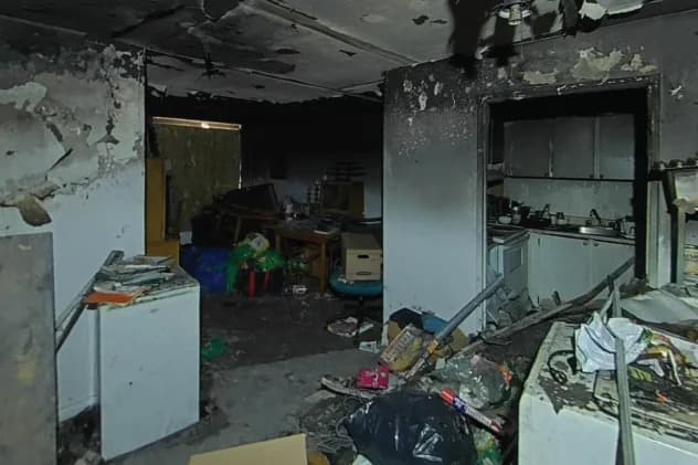 Kitchen gutted by fire, with the ceiling burned through to the framing, soot across every surface, appliances and cabinets destroyed, and debris covering the floor