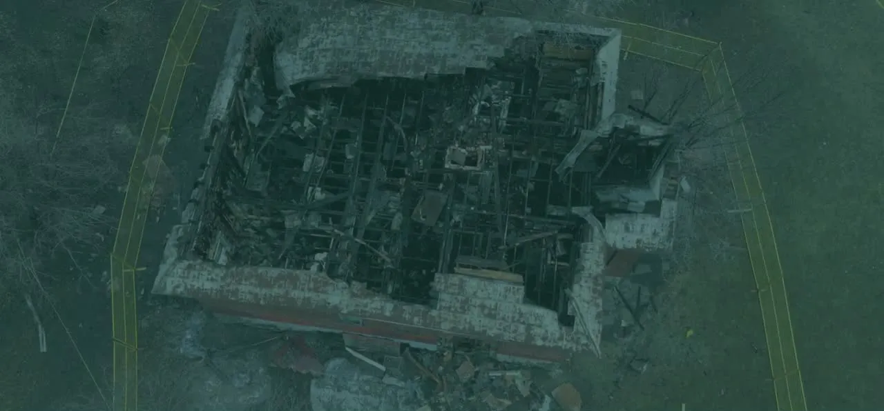 Aerial view of a fire-gutted brick building with its roof burned away, exposed floor framing and debris inside the standing walls, enclosed by temporary site fencing